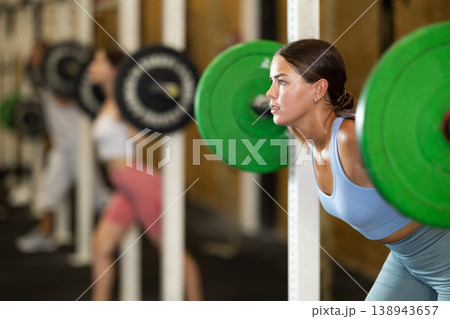 Motivated crossfit female athlete lifting heavy body bar or barbell during group training in sport gym Motivated crossfit female athlete lifting heavy body bar or barbell during group training in sport gym 138943657