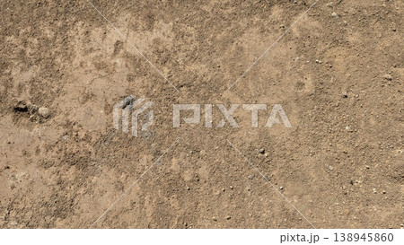 Natural dry soil surface displays a rough, grainy texture. Tiny pebbles, organic debris scatter across the parched ground. Earth tones dominate this minimalist landscape. High angle view. Natural dry soil surface displays a rough, grainy texture. Tiny pebbles, organic debris scatter across the parched ground. Earth tones dominate this minimalist landscape. High angle view. 138945860