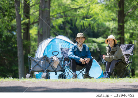 新緑のキャンプ場で椅子に座るカメラ目線笑顔のアウトドアやレジャーを楽しいミドルの夫婦 138946452