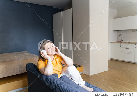 Smiling woman enjoying audio with wireless headphones, sitting comfortably on a sofa in a modern apartment. Woman relaxing at home, listening to music Smiling woman enjoying audio with wireless headphones, sitting comfortably on a sofa in a modern apartment. Woman relaxing at home, listening to music 138947484