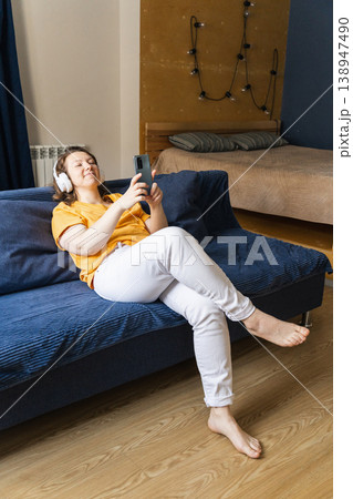 Woman wearing headphones sitting on a blue sofa, enjoying music, surfing the internet and using a smartphone in her cozy living room 138947490