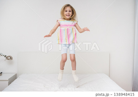 A smiling girl jumps on a bed with arms spread out in a bright minimalist room wearing a colorful rainbow striped shirt joyful energetic childhood play home family lifestyle concept A smiling girl jumps on a bed with arms spread out in a bright minimalist room wearing a colorful rainbow striped shirt joyful energetic childhood play home family lifestyle concept 138948415