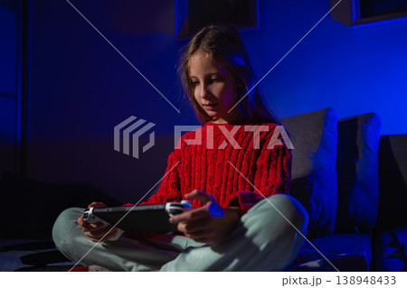 Girl sitting cross-legged on a bed looking at a handheld console screen in a dark blue-lit room Girl sitting cross-legged on a bed looking at a handheld console screen in a dark blue-lit room 138948433