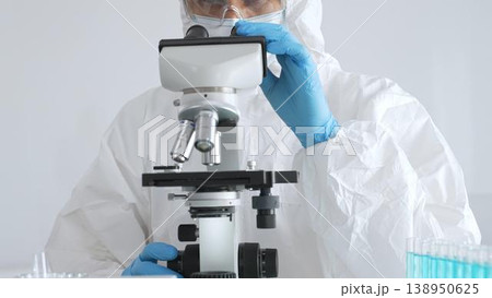 A male doctor in a protective suit and gloves uses a microscope while analyzing samples, conducting research in a laboratory. Close-up of test tubes with blue liquid A male doctor in a protective suit and gloves uses a microscope while analyzing samples, conducting research in a laboratory. Close-up of test tubes with blue liquid 138950625