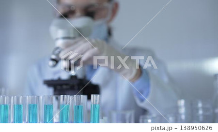 Laboratory tubes, filled with a blue liquid, are in focus, closeup view. Female scientist researcher wearing white gloves and protective mask is using a microscope at the background Laboratory tubes, filled with a blue liquid, are in focus, closeup view. Female scientist researcher wearing white gloves and protective mask is using a microscope at the background 138950626