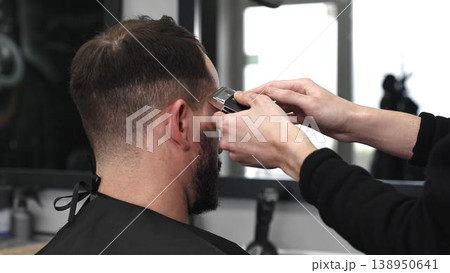 Professional barber trimming male client's hair with electric razor, delivering precise grooming service in contemporary, well lit barbershop setting Professional barber trimming male client's hair with electric razor, delivering precise grooming service in contemporary, well lit barbershop setting 138950641