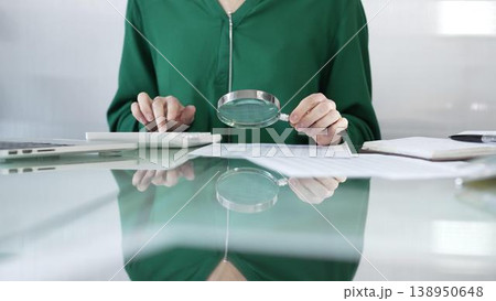 Businesswoman in green blouse carefully reviewing financial reports with magnifying glass and calculator, conducting audit and analyzing data in office. Taxes, audit in business 138950648