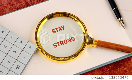 A golden magnifying glass reveals the motivating words Stay Strong written in red on a sheet of white paper A golden magnifying glass reveals the motivating words Stay Strong written in red on a sheet of white paper 138953473