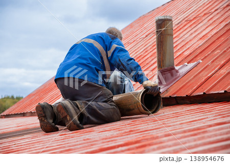 Chimney sweep worker installing pipe on red metal roof. Roofer fixing flue system on house exterior. Repair of heating ventilation and construction maintenance concept. Professional builder. Chimney sweep worker installing pipe on red metal roof. Roofer fixing flue system on house exterior. Repair of heating ventilation and construction maintenance concept. Professional builder. 138954676