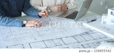 Architects engaging in a professional meeting, reviewing detailed blueprints on a table, utilizing a laptop, and collaborating on building design and construction planning 138955542