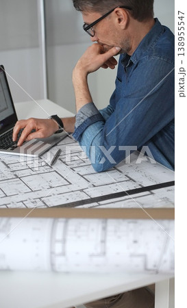 Architect man working on a laptop, analyzing blueprint plans and residential building models, concentrating on urban planning and architectural design in a modern office Architect man working on a laptop, analyzing blueprint plans and residential building models, concentrating on urban planning and architectural design in a modern office 138955547