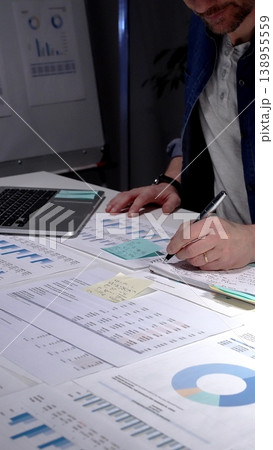 Man working late in a dim office, studying financial reports and charts across his desk while using a laptop, focused on deadlines, strategy and business growth Man working late in a dim office, studying financial reports and charts across his desk while using a laptop, focused on deadlines, strategy and business growth 138955559