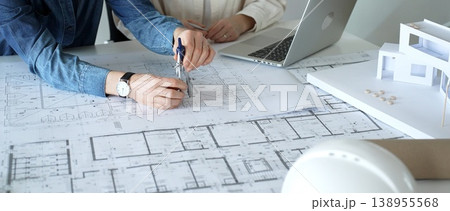 Engineering professionals collaborating on an architectural blueprint, using a compass to design a modern building, planning construction with a house model and laptop Engineering professionals collaborating on an architectural blueprint, using a compass to design a modern building, planning construction with a house model and laptop 138955568