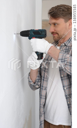 Middle aged male professional worker wearing grey checked shirt and white gloves, is drilling hole in a wall with cordless drill during home renovation work. Vertical portrait view Middle aged male professional worker wearing grey checked shirt and white gloves, is drilling hole in a wall with cordless drill during home renovation work. Vertical portrait view 138955585