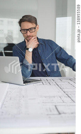 Male architect in glasses at a contemporary office working on a white architectural scale model of a modern house design, developing creative housing plans on a desk with blueprints and a laptop Male architect in glasses at a contemporary office working on a white architectural scale model of a modern house design, developing creative housing plans on a desk with blueprints and a laptop 138955589
