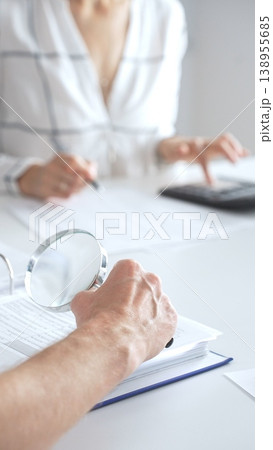 Close up of accountant hand using a magnifying glass and a calculators, to audit financial reports in brightly lit office. Audit and taxes concept 138955685