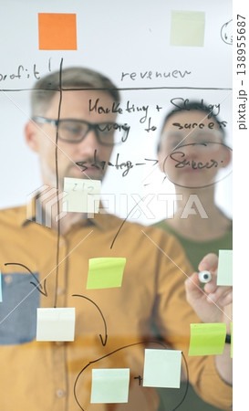 Business people colleagues planning strategy, writing notes on glass wall, collaborating with enthusiasm in bright contemporary office workspace 138955687