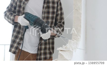 Close up of unrecognizable male construction worker wearing beige checkered shirt and protective gloves, is demolishing white wall with rotary hammer drill, generating dust, close up view. Renovation 138955709