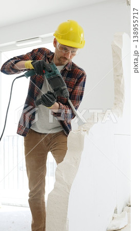 Male construction worker wearing red checkered shirt, yellow protective helmet, is demolishing white wall with rotary hammer drill, generating dust, medium long shot. Renovation project Male construction worker wearing red checkered shirt, yellow protective helmet, is demolishing white wall with rotary hammer drill, generating dust, medium long shot. Renovation project 138955717