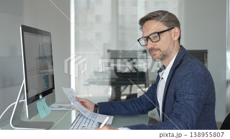 Professional man in a blue jacket working at office desk, analyzing documents and using computer for business productivity. Business people concept Professional man in a blue jacket working at office desk, analyzing documents and using computer for business productivity. Business people concept 138955807