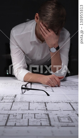 Architect covering his face with his hand, showing stress and burnout while working on complex blueprints late at night. Concept of architecture and business in low key lighting Architect covering his face with his hand, showing stress and burnout while working on complex blueprints late at night. Concept of architecture and business in low key lighting 138955813