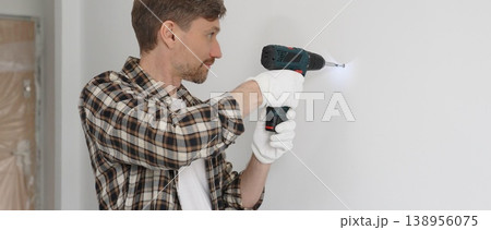 Middle aged man wearing checked shirt and white gloves, is drilling hole in a wall with cordless drill during home renovation work. Portrait view 138956075