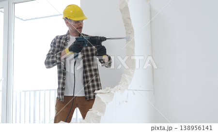 Male construction worker wearing beige checkered shirt, yellow hard hat and protective gloves, is demolishing white wall with rotary hammer drill, generating dust, medium long shot. Renovation Male construction worker wearing beige checkered shirt, yellow hard hat and protective gloves, is demolishing white wall with rotary hammer drill, generating dust, medium long shot. Renovation 138956141