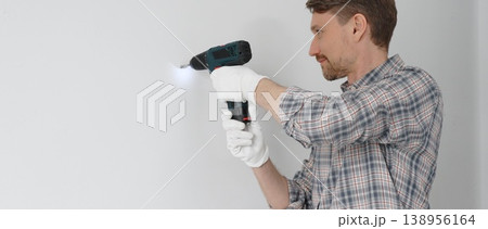 Middle aged male professional worker wearing grey checked shirt and white gloves, is drilling hole in a wall with cordless drill during home renovation work. Horizontal portrait view 138956164