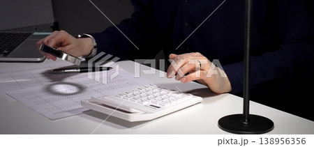 Person checking financial documents and numbers with a magnifying glass, analyzing business data for budgeting and taxes at night. Audit and taxes in business Person checking financial documents and numbers with a magnifying glass, analyzing business data for budgeting and taxes at night. Audit and taxes in business 138956356