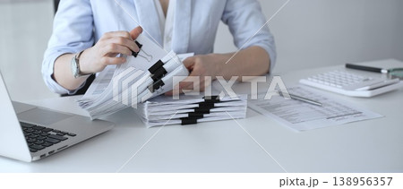 Businesswoman managing heavy workload, organizing financial documents for corporate accounting, tax preparation, and audit, working at office desk with stack of paper files and laptop. Audit and taxes Businesswoman managing heavy workload, organizing financial documents for corporate accounting, tax preparation, and audit, working at office desk with stack of paper files and laptop. Audit and taxes 138956357