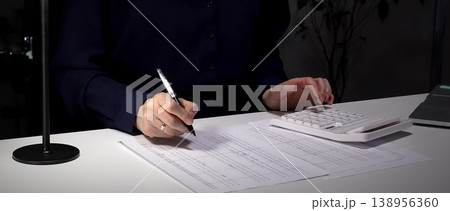 Woman calculating numbers on calculator, reviewing financial documents, working late in dimly lit office. Audit and taxes in business Woman calculating numbers on calculator, reviewing financial documents, working late in dimly lit office. Audit and taxes in business 138956360
