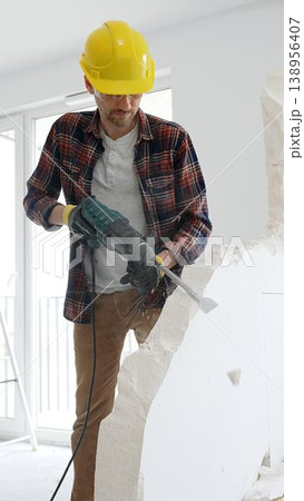 Man construction worker wearing red checkered shirt , yellow hard hat and protective gloves, is demolishing white wall with rotary hammer drill, generating dust, medium long shot. Renovation concept Man construction worker wearing red checkered shirt , yellow hard hat and protective gloves, is demolishing white wall with rotary hammer drill, generating dust, medium long shot. Renovation concept 138956407