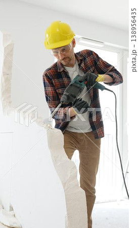 Male construction worker wearing red checkered shirt, yellow protective helmet, is demolishing white wall with rotary hammer drill, generating dust, medium long shot. Renovation project Male construction worker wearing red checkered shirt, yellow protective helmet, is demolishing white wall with rotary hammer drill, generating dust, medium long shot. Renovation project 138956435