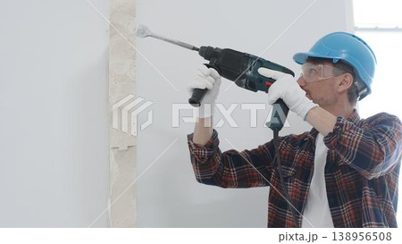 Male construction worker wearing red checkered shirt, blue protective helmet and gloves, is demolishing white wall with rotary hammer drill, generating dust, portrait view. Renovation concept Male construction worker wearing red checkered shirt, blue protective helmet and gloves, is demolishing white wall with rotary hammer drill, generating dust, portrait view. Renovation concept 138956508