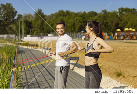 Happy couple doing stretching exercises with rubber band standing in the park outdoors Happy couple doing stretching exercises with rubber band standing in the park outdoors 138956509