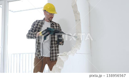 Male construction worker wearing beige checkered shirt, yellow hard hat and protective gloves, is demolishing white wall with rotary hammer drill, generating dust, medium long shot. Renovation project Male construction worker wearing beige checkered shirt, yellow hard hat and protective gloves, is demolishing white wall with rotary hammer drill, generating dust, medium long shot. Renovation project 138956567