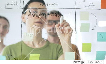 Professional businesswoman wearing glasses writing on glass board, marking strategic plan with sticky notes while colleagues brainstorming in background 138956598