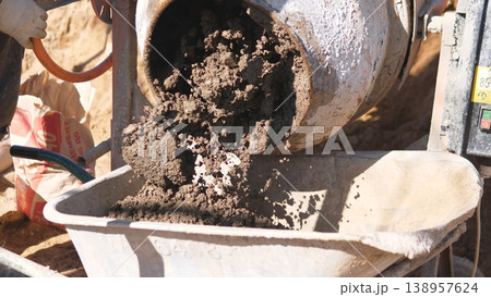 Concrete Pouring From Mixer Into Wheelbarrow Via Chute, Wet Flow And Granular Texture Visible, Operator Controlling Discharge, Work Boots And Site Tools Nearby, Efficient Material Transfer Concrete Pouring From Mixer Into Wheelbarrow Via Chute, Wet Flow And Granular Texture Visible, Operator Controlling Discharge, Work Boots And Site Tools Nearby, Efficient Material Transfer 138957624