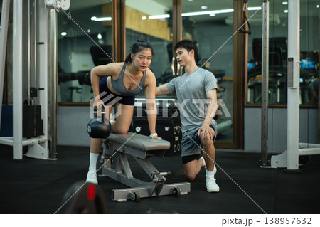 A woman is performing a kettlebell row on a bench while her trainer closely checks her form A woman is performing a kettlebell row on a bench while her trainer closely checks her form 138957632