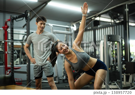 A woman is stretching and warming up at the gym while her trainer supervises and encourages her A woman is stretching and warming up at the gym while her trainer supervises and encourages her 138957720