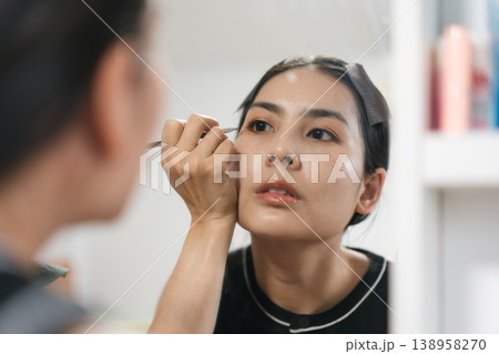 Beautiful Thai woman applying makeup with eye liner, eyebrow pencil in front of mirror. Lifestyle concept for cosmetics and self care 138958270