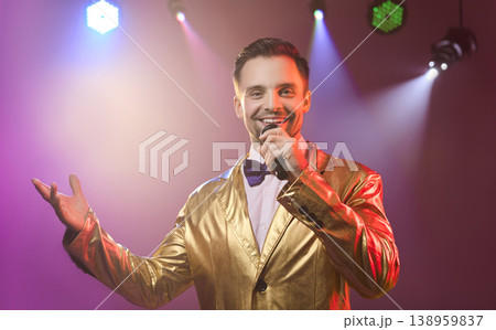 Smiling showman in gold suit singing with microphone under disco lights on stage Smiling showman in gold suit singing with microphone under disco lights on stage 138959837