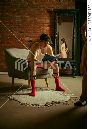 Man reading book in armchair ignoring woman reflected in mirror in loft interior. 138961180