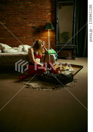 Woman sitting by open suitcase with phone in hand in moody loft bedroom interior. 138961186