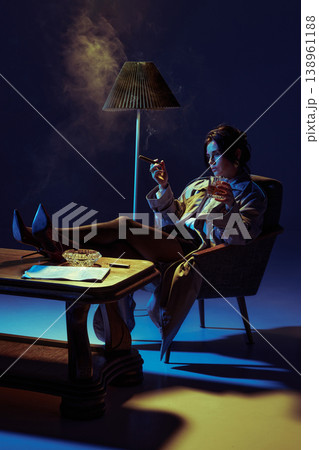 Elegant woman smoking cigar and drinking whiskey in armchair under dramatic studio lighting. 138961188