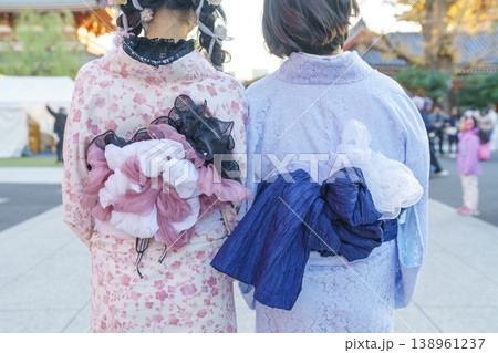 Rear view of two women in elegant pink and blue floral kimonos with elaborate, decorative obi bows at a Japanese temple. Highlighting traditional craftsmanship and fashion in a grand cultural setting. 138961237