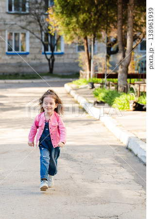 Little girl walking alone on residential street wearing pink jacket and denim overalls during daytime in urban neighborhood. Concept of childhood independence lifestyle movement and outdoor activity 138961939