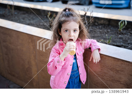 Little girl eating ice cream cone outdoors standing near urban planter and looking at camera with curious expression. Concept of childhood food enjoyment and casual lifestyle Little girl eating ice cream cone outdoors standing near urban planter and looking at camera with curious expression. Concept of childhood food enjoyment and casual lifestyle 138961943