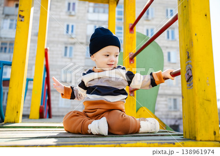 Infant child sitting on playground platform holding metal bar and looking aside with curious calm expression in urban yard. Concept of childhood development activity and exploration 138961974
