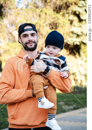 Father holding toddler boy in arms outdoors in park while both look at camera and child points forward. Concept of parenting and family care Father holding toddler boy in arms outdoors in park while both look at camera and child points forward. Concept of parenting and family care 138962051